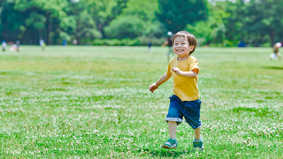 芝生の上を笑顔で走っている男の子。黄色いTシャツを着てデニムのハーフパンツを履いて、カメラに向かって走ってきている。