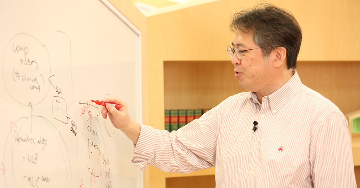 眼鏡をかけた男性がホワイトボードを指しながら説明している場面。
