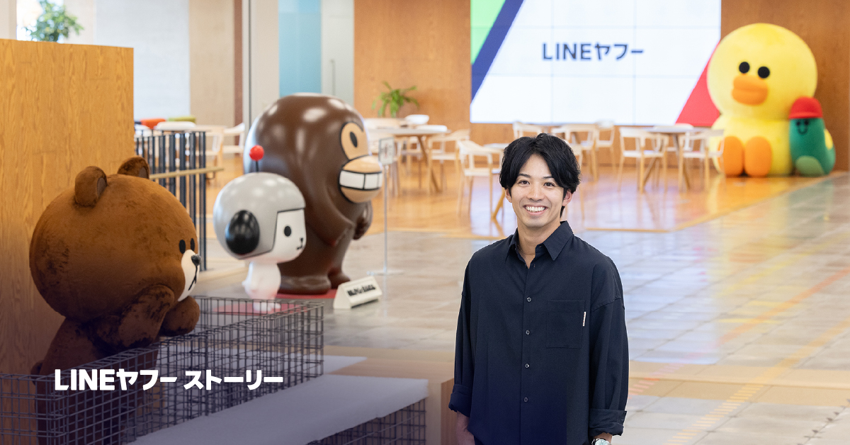 LINEヤフーのオフィスで、けんさくとえんじん、ブラウン、サリーなどキャラクターのぬいぐるみが並ぶ中、笑顔の男性、益田が立っている写真です。