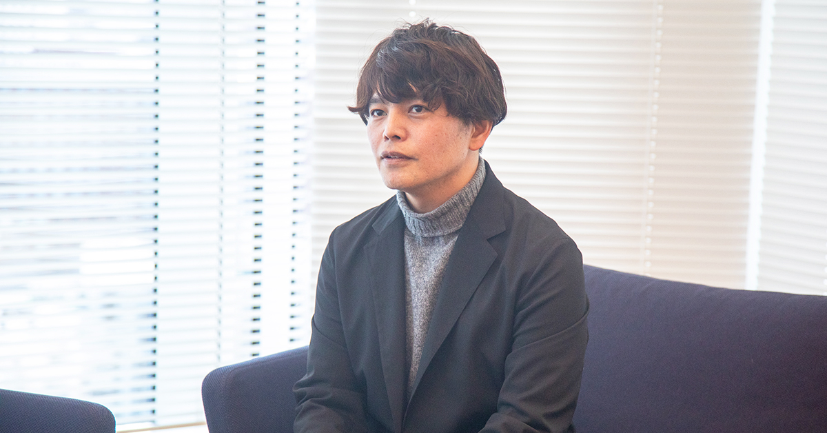 男性がソファに座り、真剣な表情で話をしています。背景はブラインドの窓。