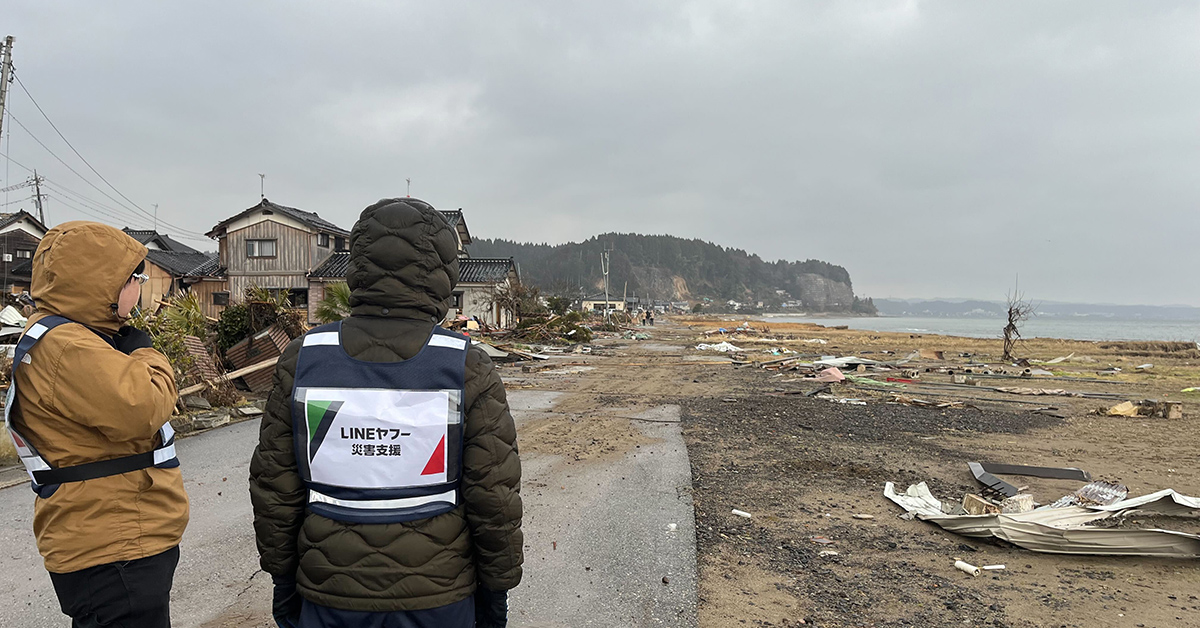 災害支援のベストを着た二人が、被災地を見渡しています。周囲は瓦礫が散乱。