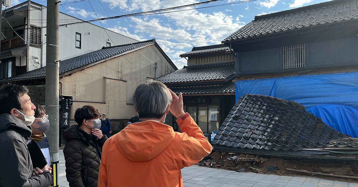 数人が古い建物を見上げています。屋根は青いシートで覆われています。