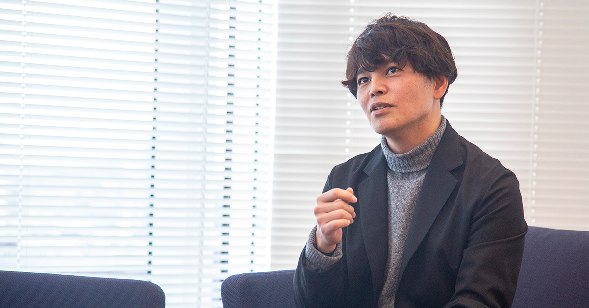 男性がソファに座り、考えながら話しています。背景はブラインドの窓。