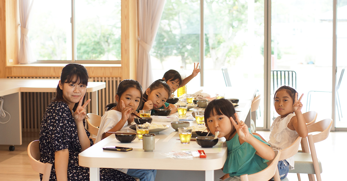 女性と子供たちが食卓で食事中。皆ピースサインをしています。