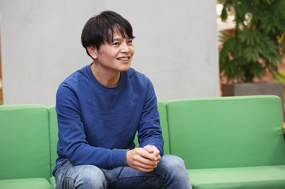 青いセーターを着た男性が緑のソファに座って微笑んでいる。リラックスした雰囲気。