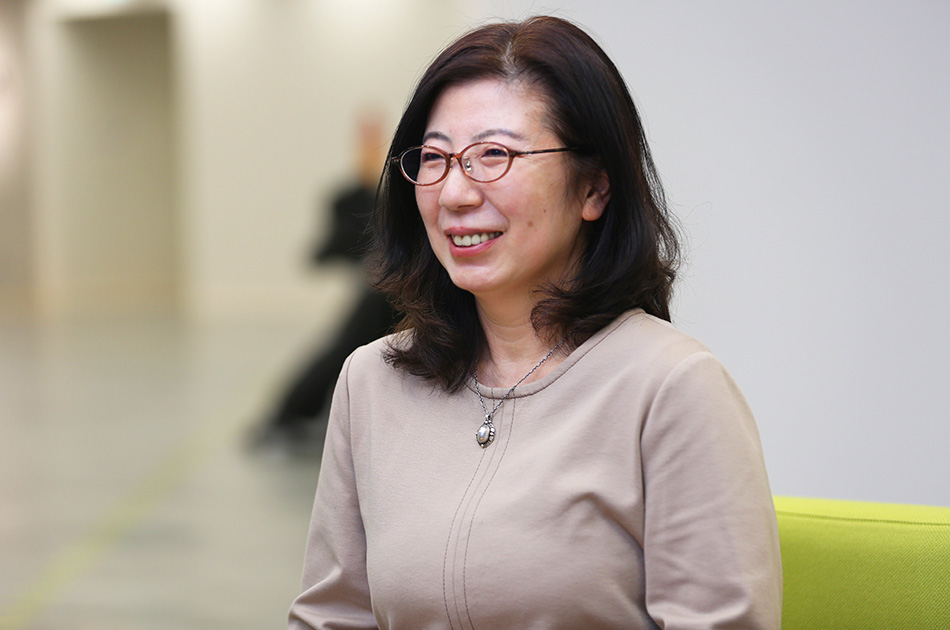 眼鏡をかけた女性が微笑んでいる。ベージュのトップスを着て、落ち着いた雰囲気。