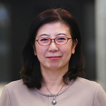 眼鏡をかけた女性がカメラに向かって微笑んでいる。落ち着いた色合いのトップスを着用。