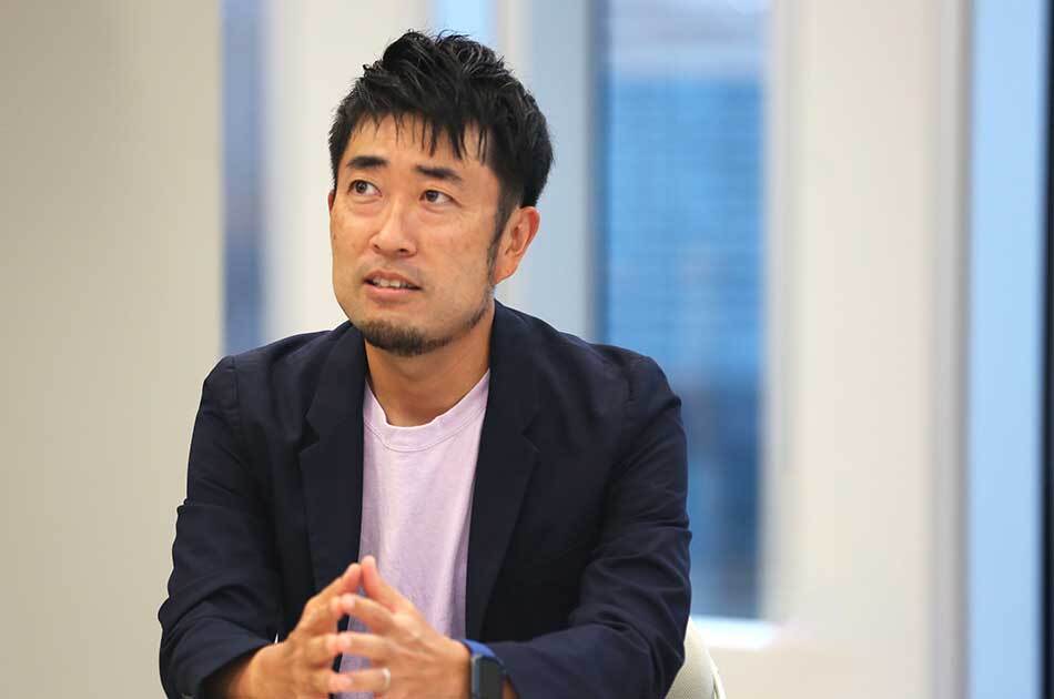 男性がインタビュー中の様子。背景には窓があり、手を組んで話している。
