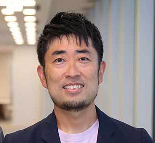 男性のポートレート写真。明るい表情で、背景にはオフィスの照明が見える。
