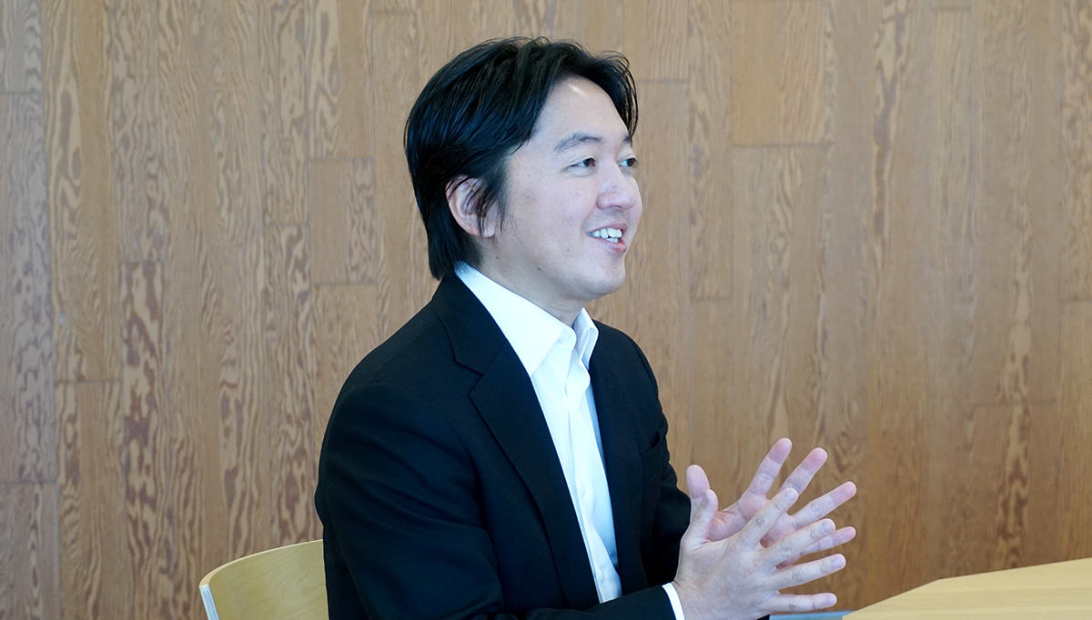 スーツ姿の男性がテーブルに座り、手を組んで笑顔で話している。