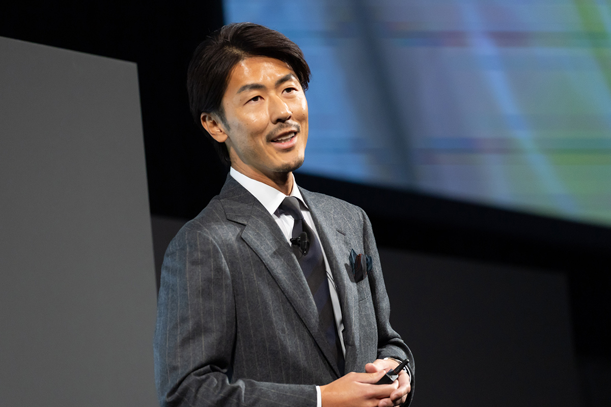 スーツを着た男性がステージでプレゼンテーションを行っている様子です。