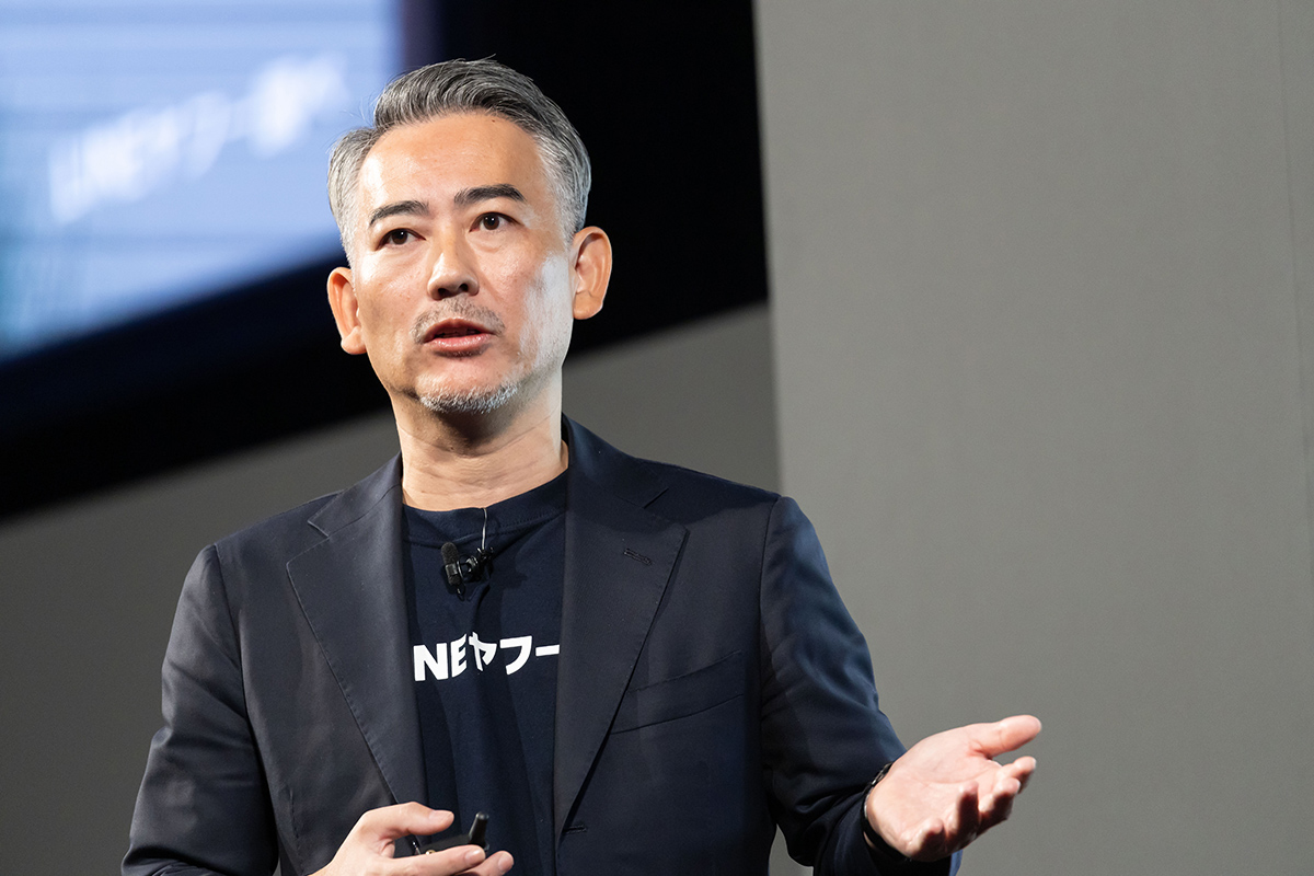 スーツを着た男性がステージで話をしている様子で、手を動かしながら説明しています。