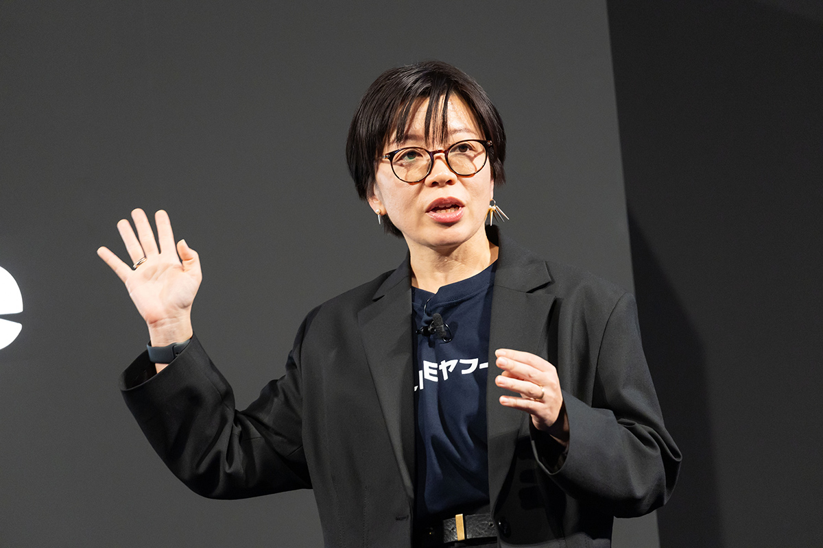 眼鏡をかけた女性がステージでプレゼンテーションを行っている様子です。