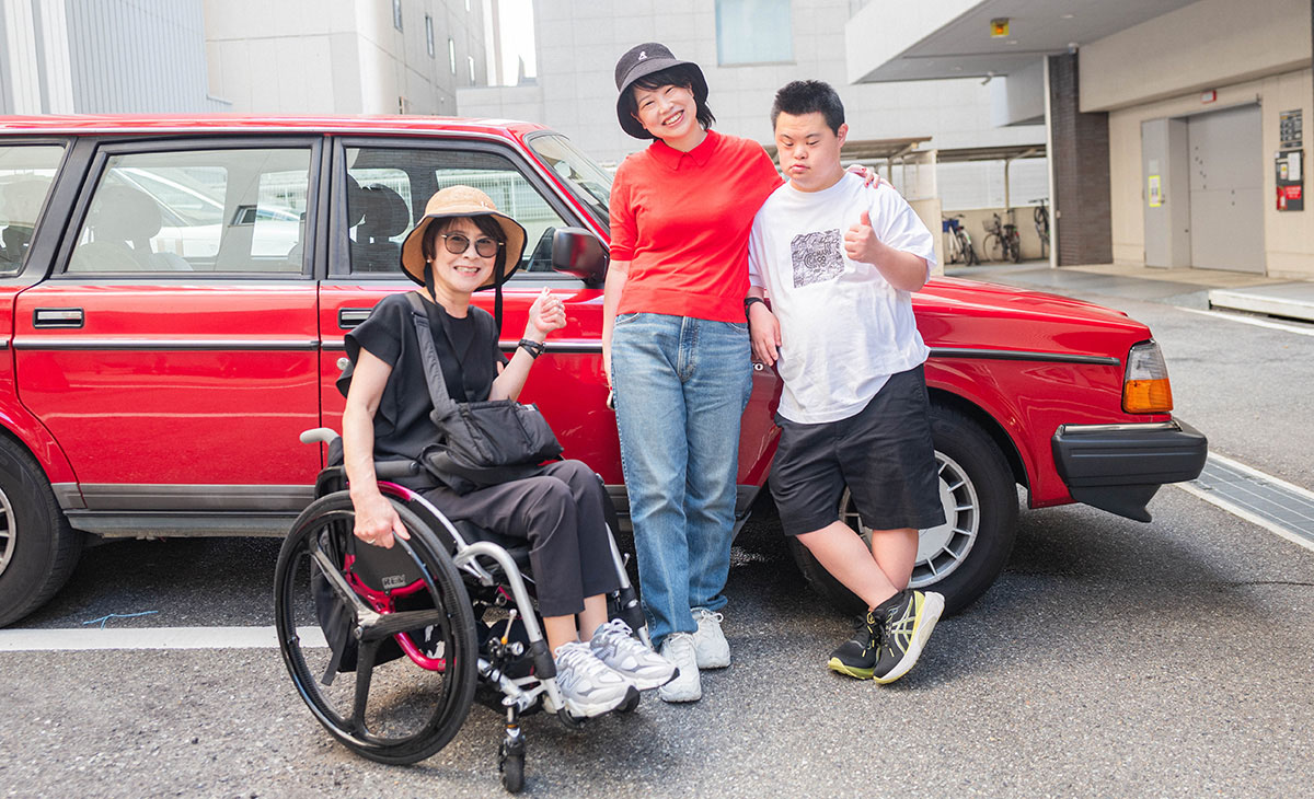 赤い車の前で、車椅子の女性と二人の仲間が笑顔でポーズをとっている。楽しそうな雰囲気。
