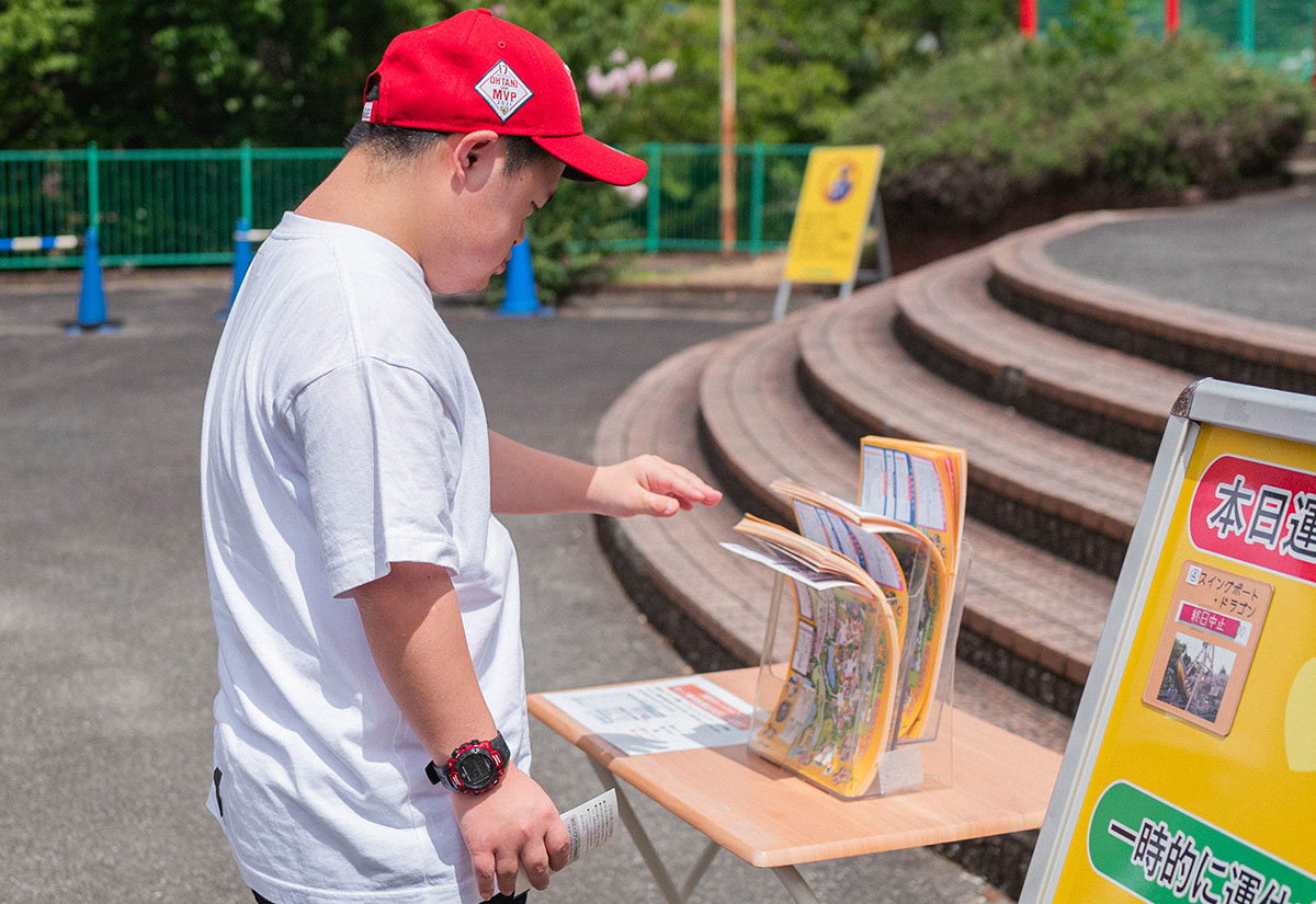 赤い帽子をかぶった少年が、案内パンフレットを手に取ろうとしている。周囲には緑が広がる。