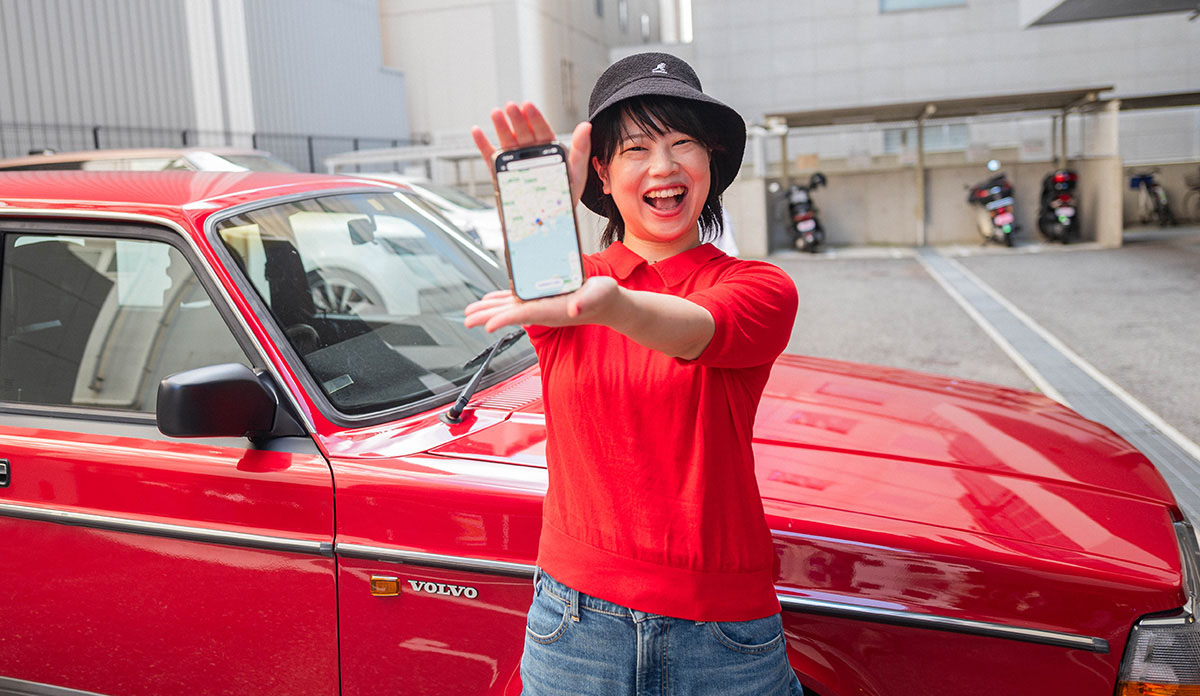 赤い車の前で女性がスマートフォンを見せて笑顔を見せている。