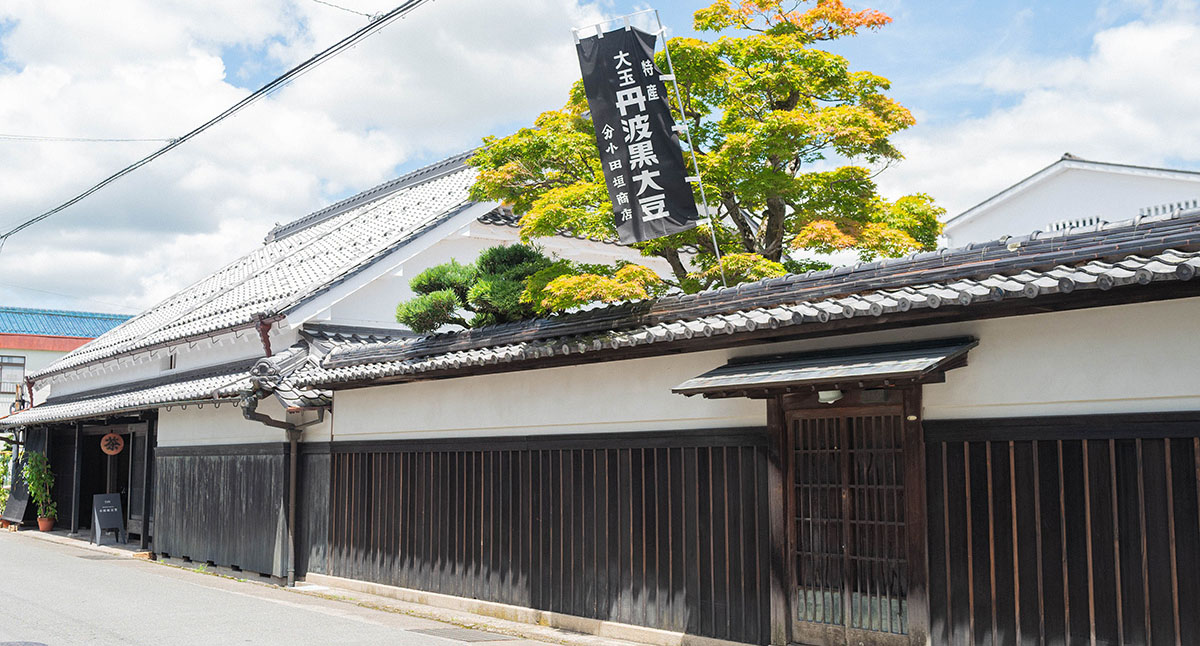 伝統的な日本家屋の外観。黒い塀と緑の木々が特徴的な景色。