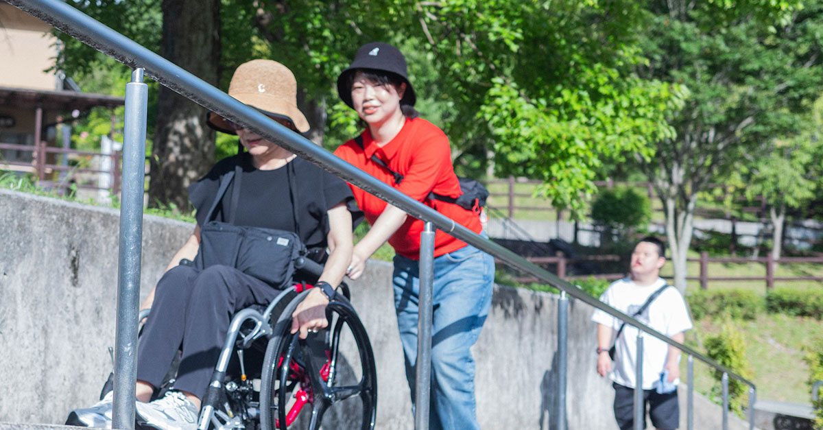 女性が車椅子に乗った人を坂道でサポートしながら押している。周囲には緑が広がっている。