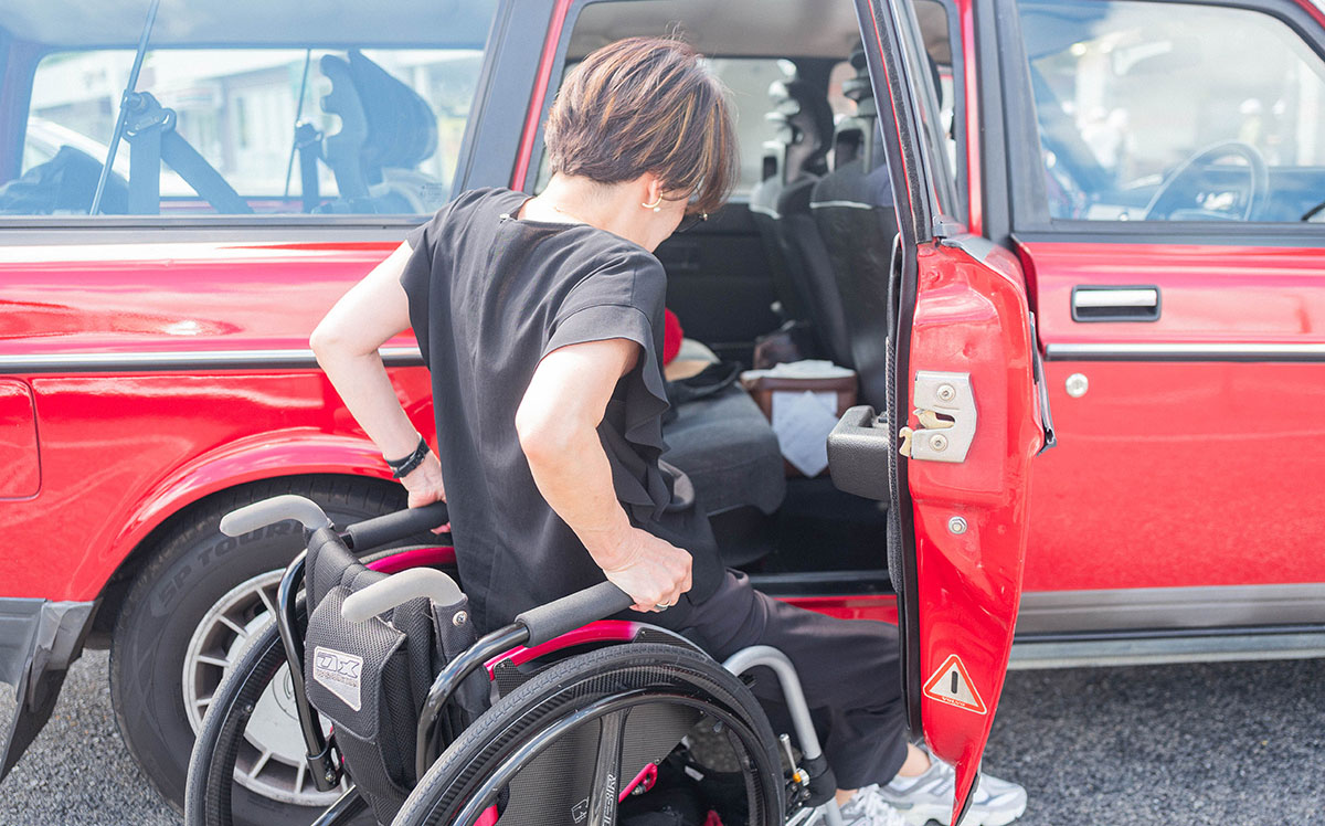 女性が車椅子から赤い車に乗り込もうとしている。周囲には明るい日差しが差し込んでいる。