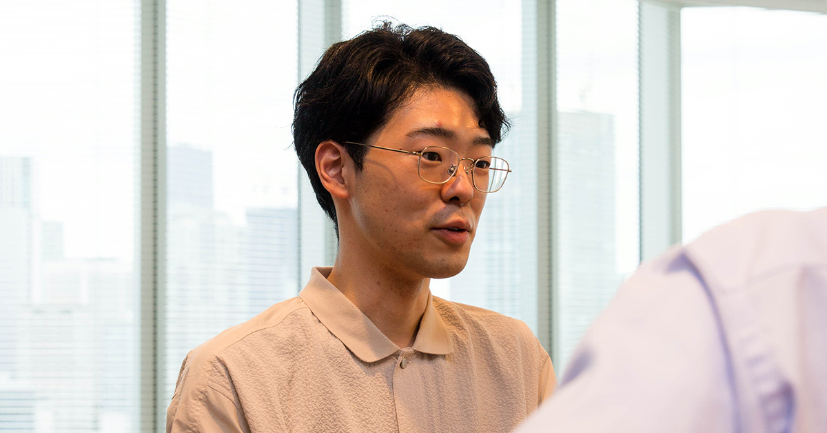 男性が会話中の様子。窓際で真剣な表情をしており、背景には都市の風景が見える。