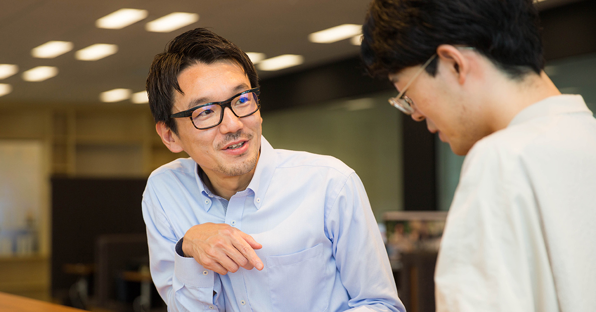 二人の男性が会話をしている。明るいオフィスで、親しげに意見を交わしている。