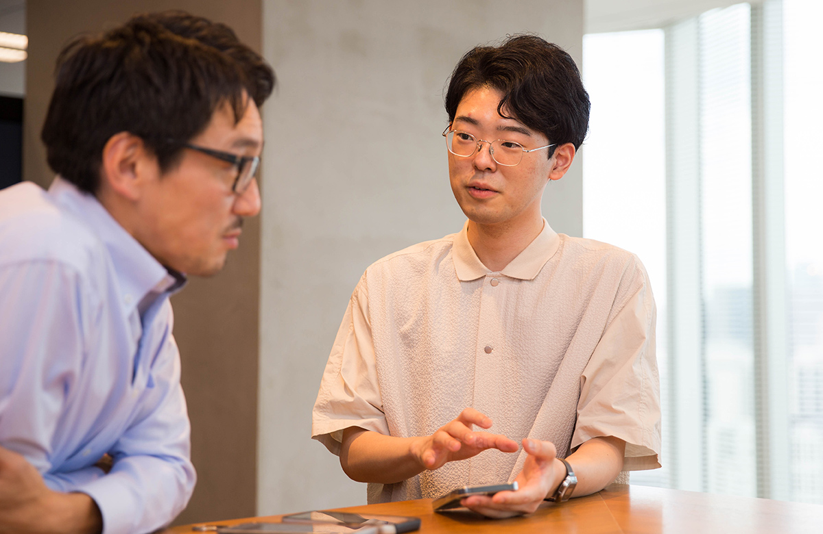 二人の男性が立ち話をしている。窓際で、スマートフォンを使いながら話し合っている。