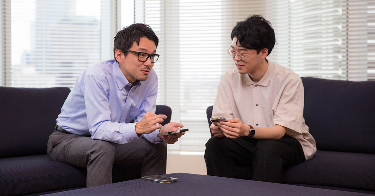 二人の男性がソファに座って会話している。スマートフォンを手に取り、親密な会話をしている。