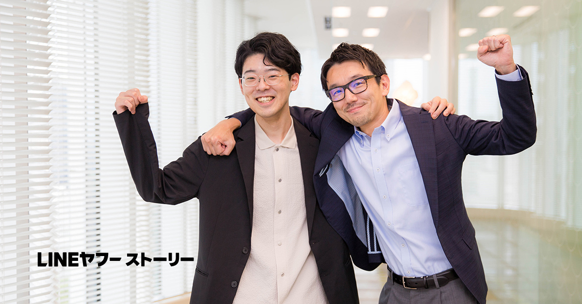 二人の男性が肩を組んでポーズをとっている。明るい廊下で、達成感に満ちた表情をしている。