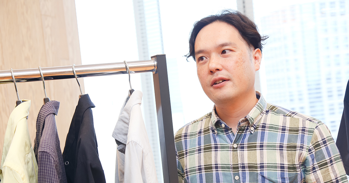 男性がチェック柄のシャツを着て、服のラックの横で話している。窓からの光が差し込み、明るい雰囲気。