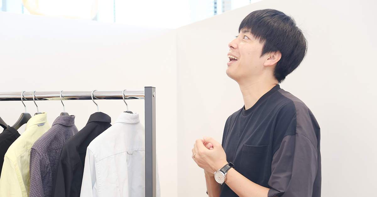 男性が笑顔で話している。腕時計をしており、ラックにかかった服の前で楽しそうにしている。