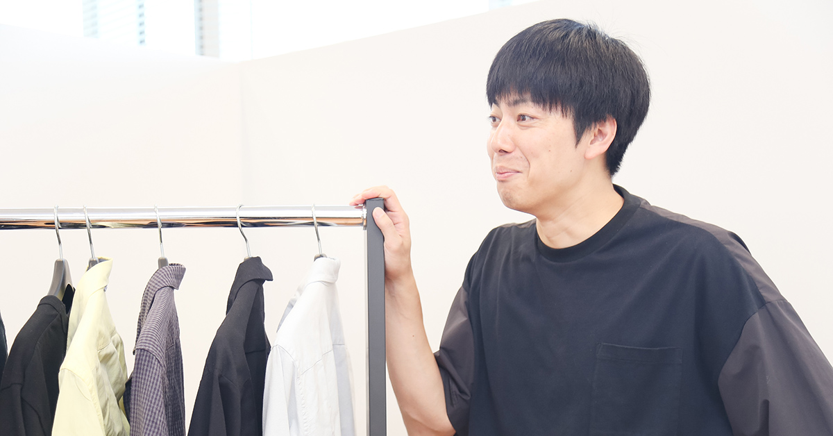 黒いシャツの男性が服のラックを片手で支えながら、微笑んでいる。リラックスした雰囲気が伝わる。