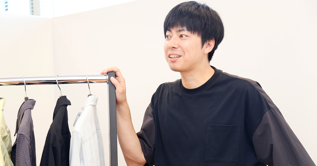 男性が服のラックの横で話している。黒いシャツを着ており、リラックスした表情で会話を楽しんでいる様子。