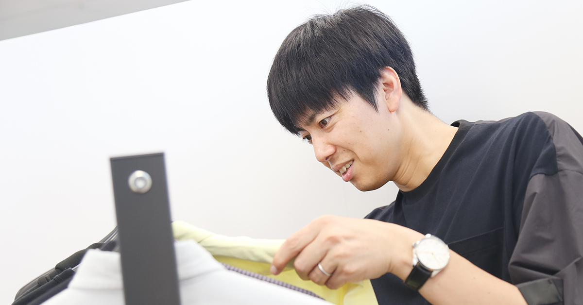 画像には、男性が服を見ている様子が写っています。彼はラックにかかった服を手に取り、興味深そうに見ています。シンプルな黒いシャツを着ており、腕時計をしています。