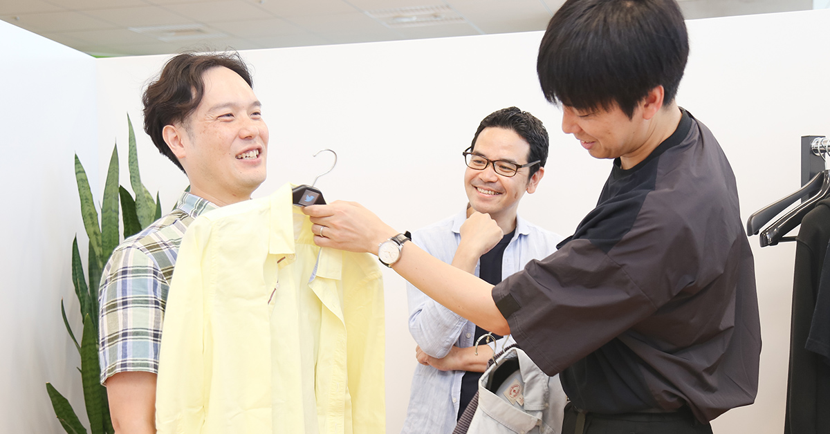 三人の男性がシャツを手に持ちながら談笑している。リラックスした雰囲気が感じられる。