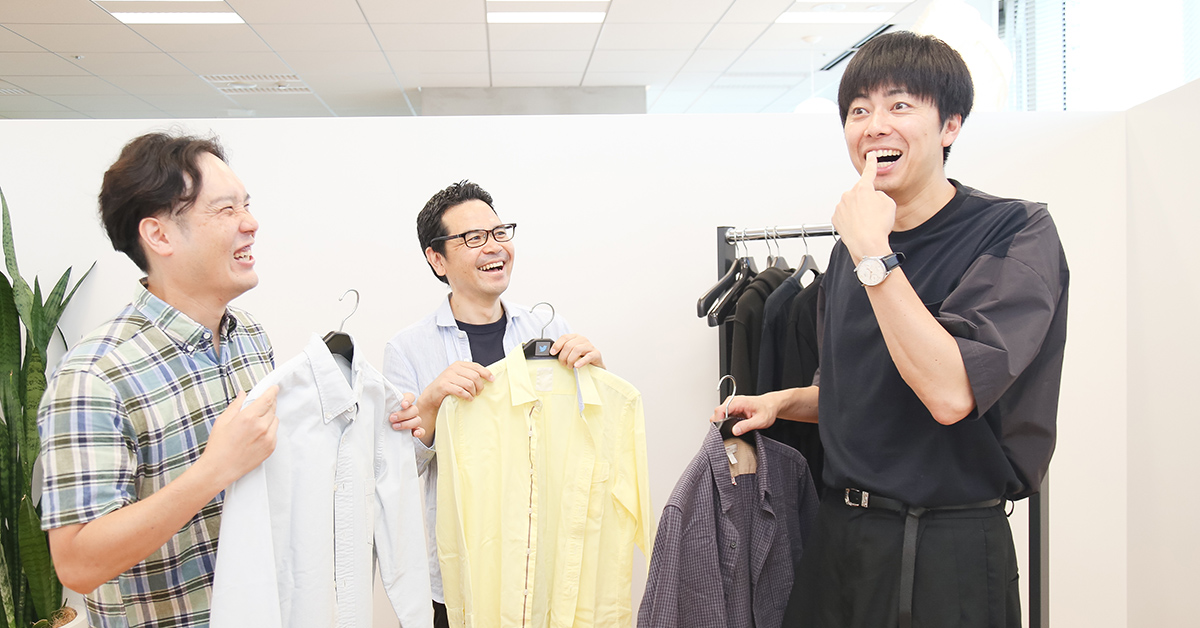 三人の男性が楽しそうにシャツを手に持ち、笑顔で会話している。明るい室内の様子。