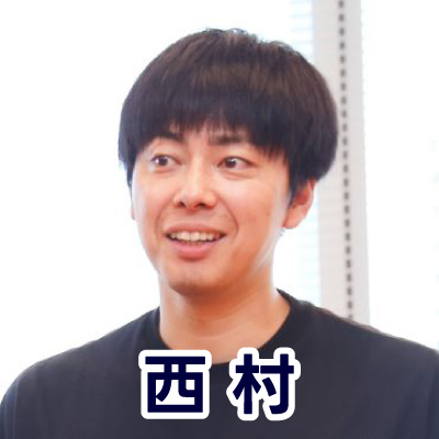 黒いシャツを着た男性がカメラに向かって笑顔を見せている。明るい室内で撮影。下部に「西村」と書かれている。