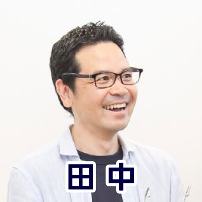 眼鏡をかけた男性が横を向いて微笑んでいる。カジュアルな服装で、落ち着いた雰囲気。下部に「田中」と書かれている。