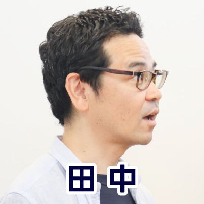 眼鏡をかけた男性が横を向いて話している。カジュアルな服装で、落ち着いた雰囲気。田中さんのアイコン。