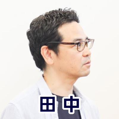 眼鏡をかけた男性が横を向いて話している。カジュアルな服装で、落ち着いた雰囲気。田中さんのアイコン。