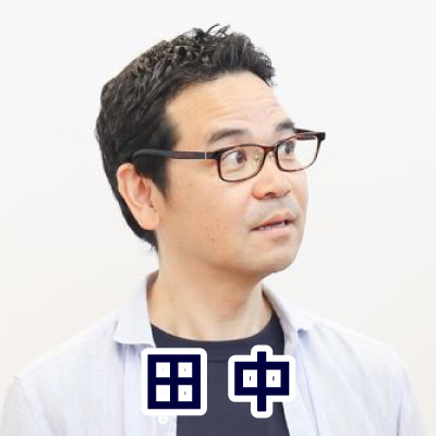 眼鏡をかけた男性が横を向いて話している。カジュアルな服装で、落ち着いた雰囲気。田中さんのアイコン。