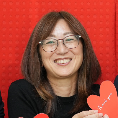 眼鏡をかけた女性が赤い背景の前で微笑んでいます。手には「Thank you」と書かれたハートを持っています。