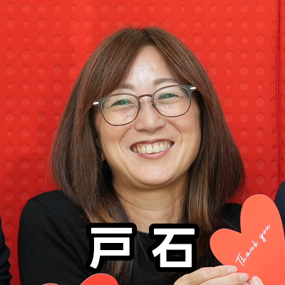 眼鏡をかけた女性が笑顔で座っており、「戸石」と書かれたテキストが重ねられています。