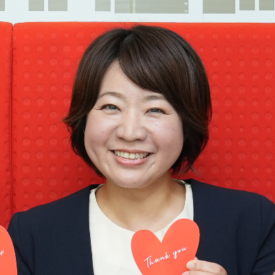 女性が赤い背景の前で微笑んでいます。手には「Thank you」と書かれたハートを持っています。