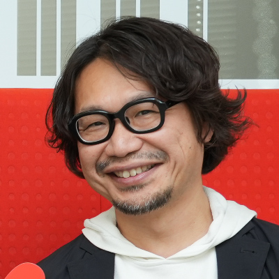 眼鏡をかけた男性が赤い背景の前で微笑んでいます。カジュアルな服装でリラックスした様子です。