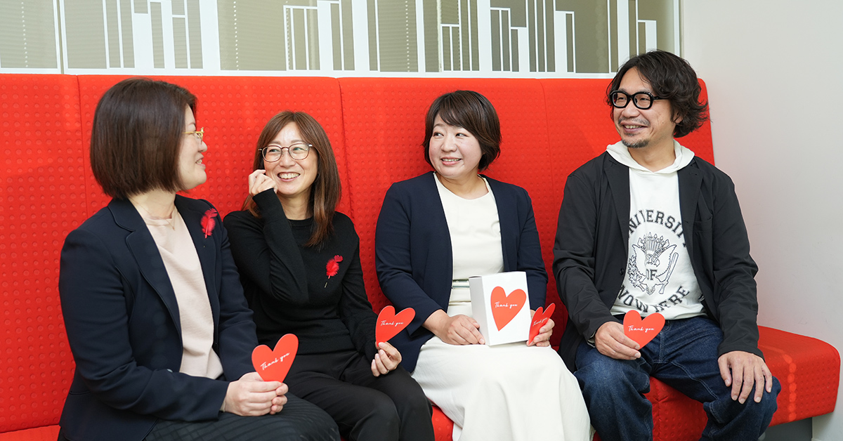 4人の人々が赤いソファに座り、手にハート型のカードを持ちながら笑顔で会話しています。