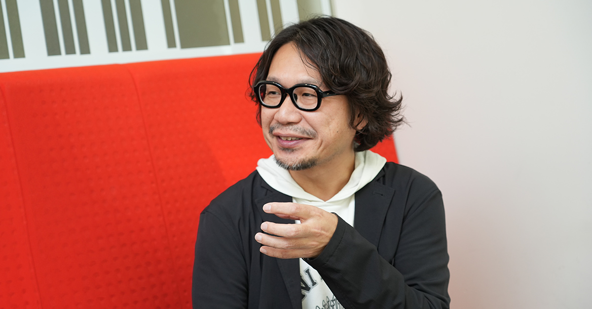 男性が赤いソファに座り、眼鏡をかけて談笑している様子が写っています。