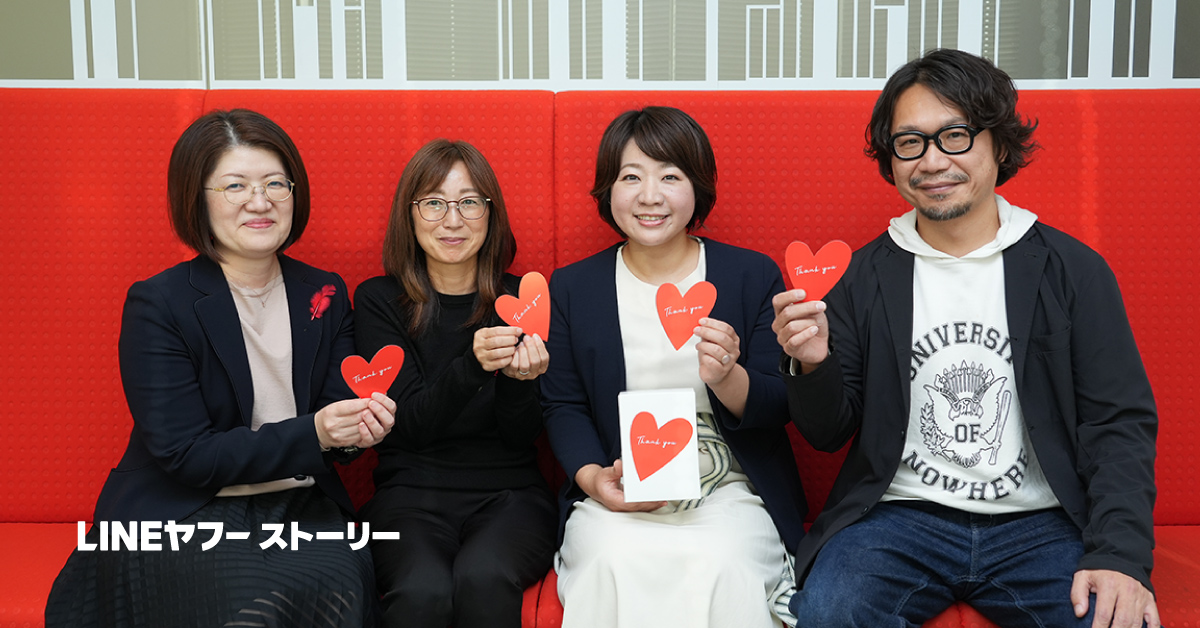 4人が赤いソファに座り、ハート型のカードを持ちながらカメラに向かって笑顔を見せています。