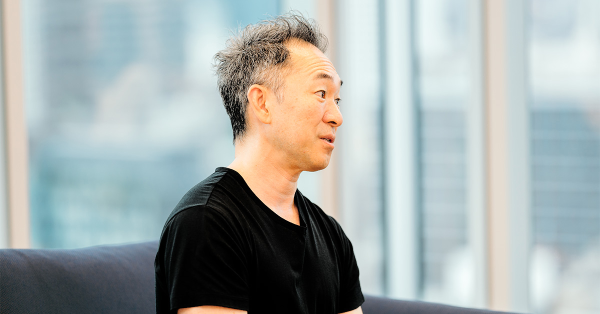 男性がソファに座り、横を向いて話をしている。背景には窓があり、都市の景色が見える。