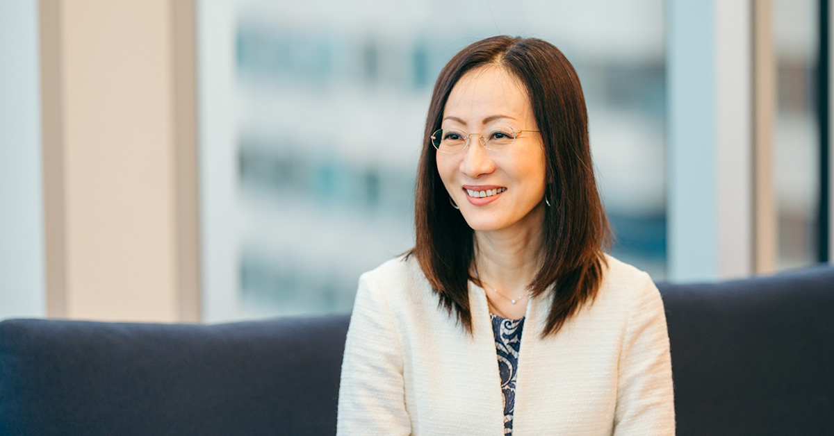 女性がソファに座り、微笑んでいる。背景には大きな窓があり、都会的な景色が広がっている。