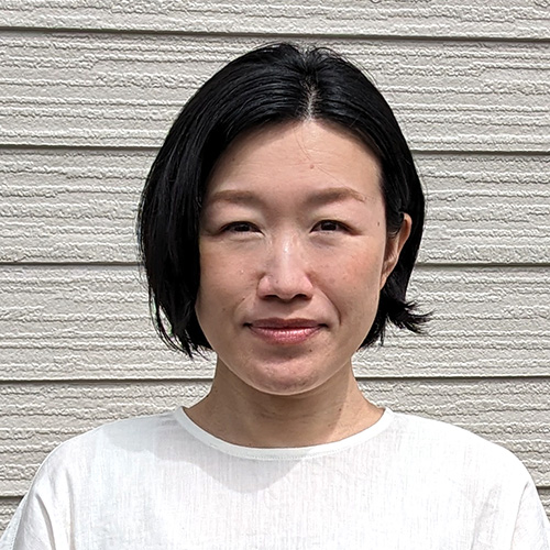 女性が白い服を着てカメラを見つめています。背景は木目調の壁で、シンプルで落ち着いた印象です。